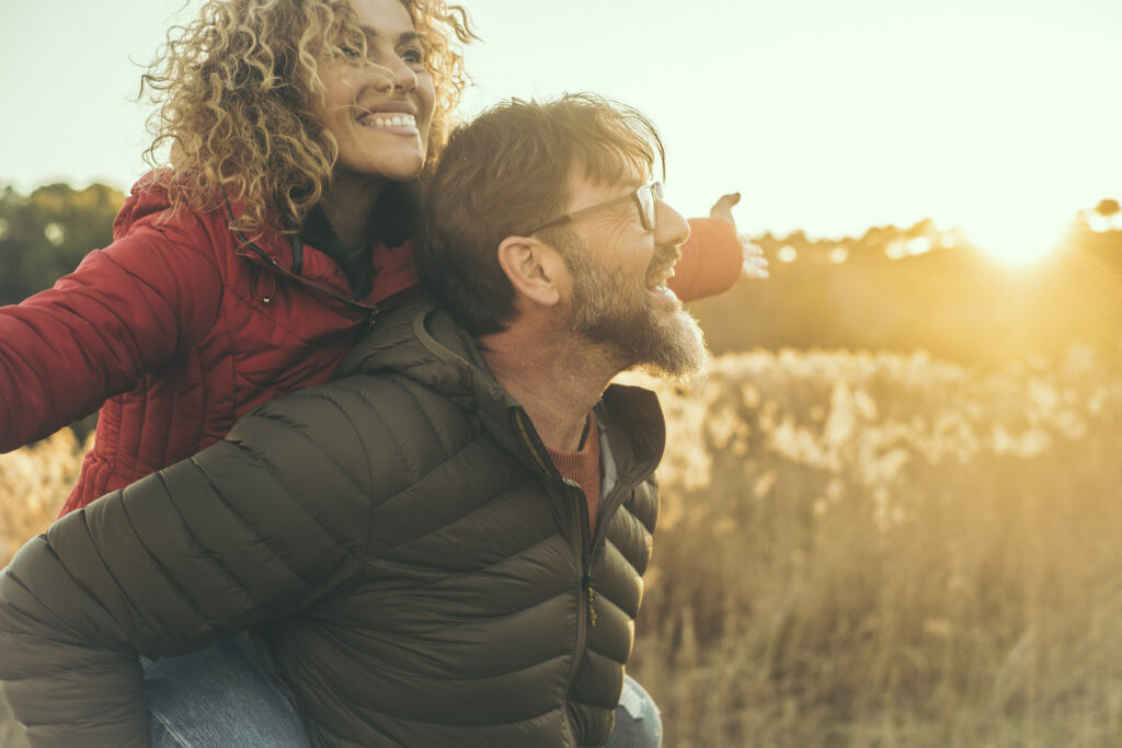 Happy couple have fun together in outdoor leisure activity in nature field during sunset time and golden hours light. Man carry woman on his back. People enjoying life and laughing. Winter autumn day