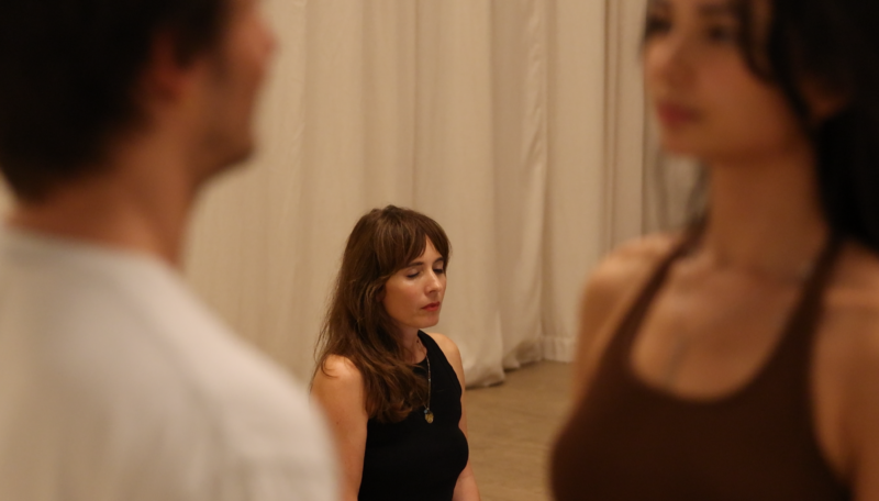 Woman instructor in black sleeveless top leading a couples workshop, with participants visible in soft focus in foreground, in warm-lit intimate space
