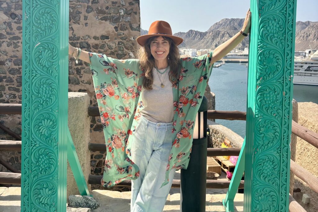 Woman standing with arms wide open in a turquoise doorway in Muscat Old Town, embodying joy and glimmers as nervous system medicine