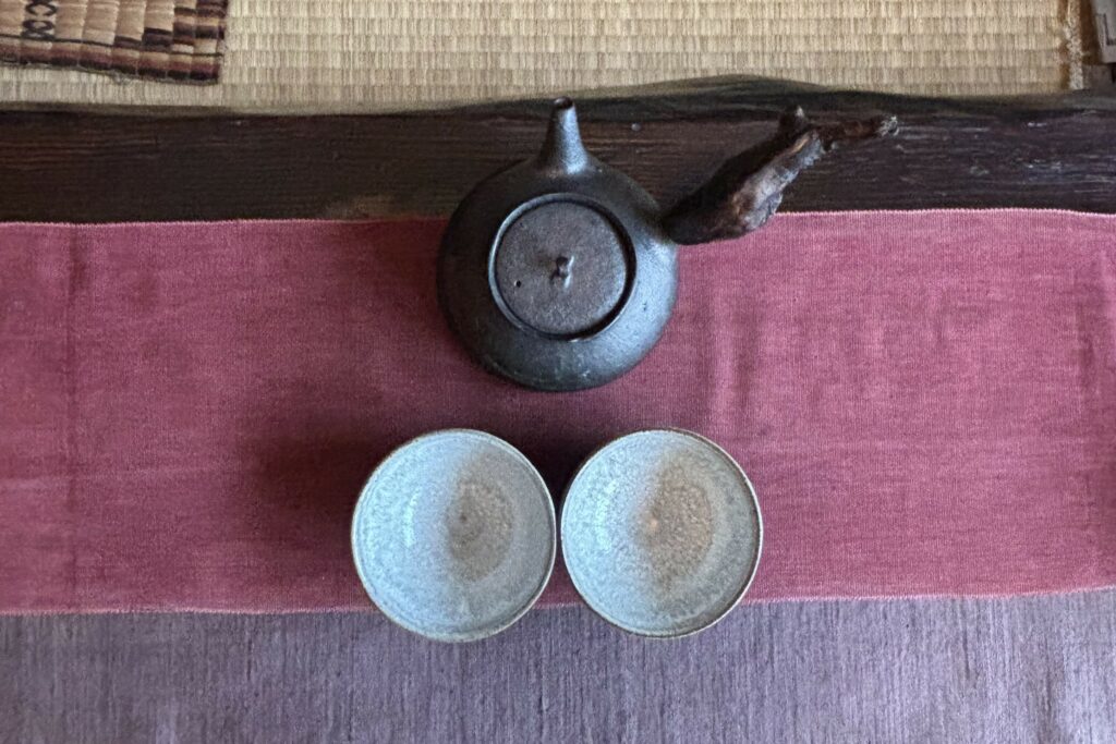 Handcrafted ceramic tea cups and dark clay teapot on a pink and mauve textile runner - the tea ceremony in Hong Kong that became one of Corinne's glimmers during a time of war and uncertainty